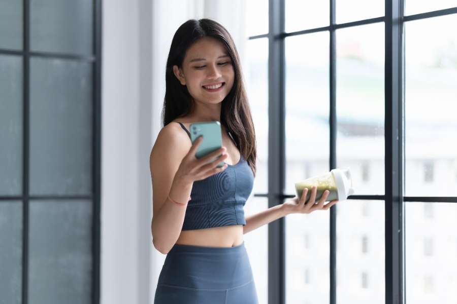 Une femme en tenue de sport consulte son téléphone en tenant un smoothie vert, suivant son régime minceur avec application.