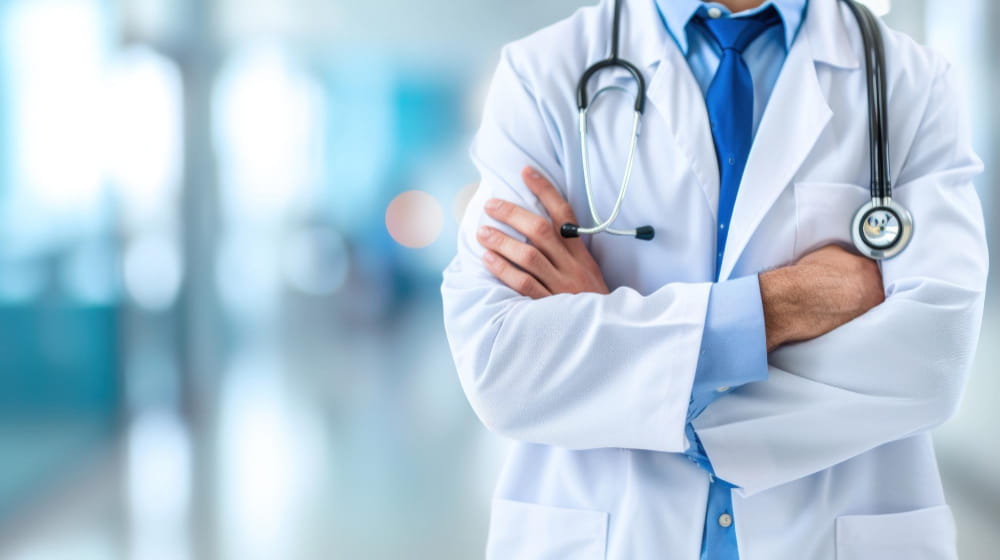 Gros plan sur un docteur en blouse blanche, bras croisés, dans le hall flou d'un hôpital moderne