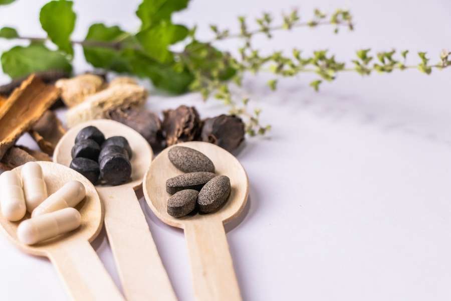 Des gélules blanches et des pilules sombres disposées sur des cuillères en bois, illustrant des compléments pour rituels ayurvédiques.