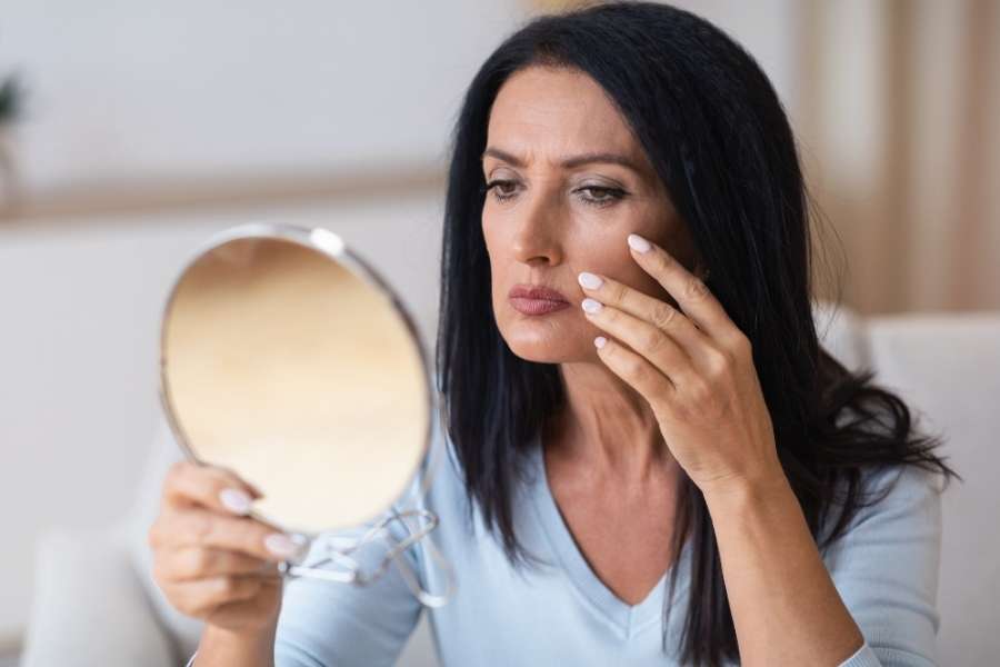 Une femme mature, l'air préoccupé, examine les signes de fatigue et de vieillissement sur sa peau dans un miroir rond.